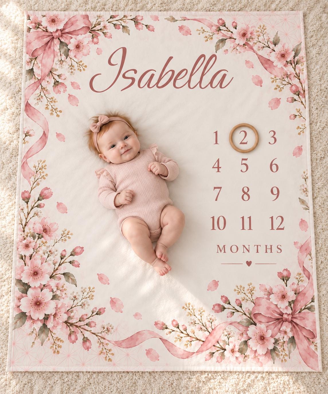 Personalized baby milestone blanket with pink cherry blossoms, ribbon bows, the sample name Isabella and months 1 to 12, with a baby lying on it and a wooden ring marking month 2, a custom baby shower gift.